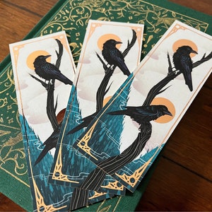 Puede incluir: Cuatro marcapáginas con un cuervo negro posado en una rama sobre un fondo azul y dorado. El cuervo está silueteado contra un sol amarillo.