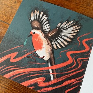 May include: A red robin with a blue teardrop flying against a dark green background with red, wavy lines. The robin has its wings spread wide and is in mid-flight.