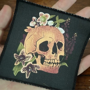 May include: A black fabric patch with a detailed illustration of a human skull surrounded by white, pink, and purple flowers. The skull is a light brown color with a detailed design.