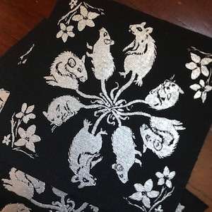 May include: A black fabric patch with a silver design of seven mice in a circle around a central point. The mice are stylized and have long tails. The background is black and the design is silver.