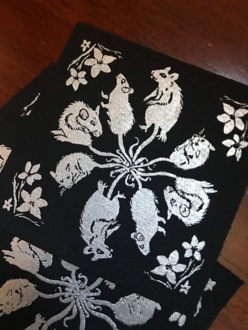 May include: A black fabric patch with a silver design of seven mice in a circle around a central point. The mice are stylized and have long tails. The background is black and the design is silver.