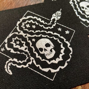 May include: Black fabric patches with a white snake and skull design. The snake is coiled around a skull, with floral and star accents. The patches are square and have a textured appearance.
