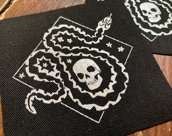 Op de afbeelding: Zwarte stoffen patches met een wit slang- en schedelontwerp. De slang kronkelt rond een schedel, met bloemen en sterren accenten. De patches zijn vierkant en hebben een getextureerde uitstraling.