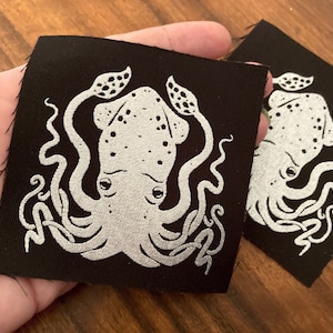 May include: A black fabric patch with a white embroidered image of a squid with tentacles. The squid has a speckled pattern and large eyes.