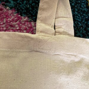 May include: Close-up of the top of a white canvas tote bag with two straps. The straps are sewn together at the top with a small, stitched X.