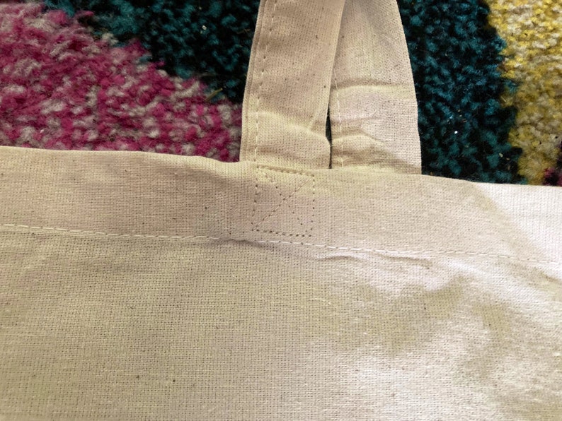 May include: Close-up of the top of a white canvas tote bag with two straps. The straps are sewn together at the top with a small, stitched X.