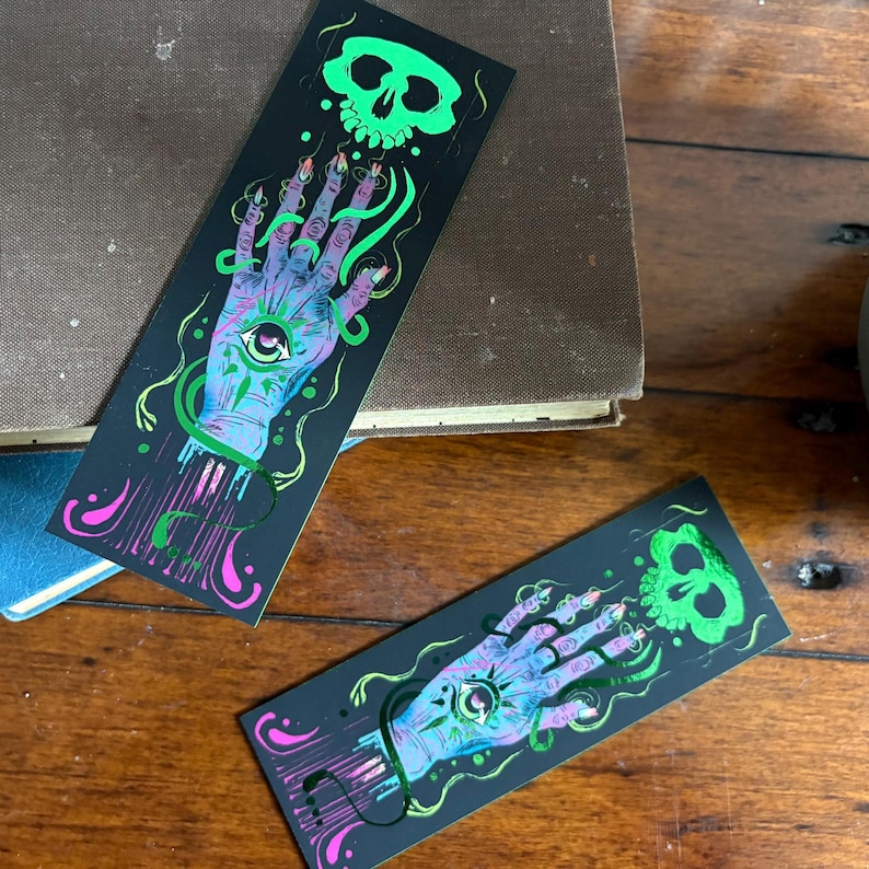 May include: Two black bookmarks with a mystical design. Each bookmark features a hand with an eye in the palm, a green skull, and green and pink accents. The hand is blue and purple, with long, pointed fingernails.