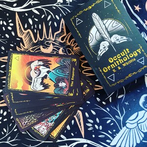May include: A set of tarot cards with black backgrounds and colorful illustrations. The cards feature a bird theme and the text "Occult Ornithology K.S. Whittle".