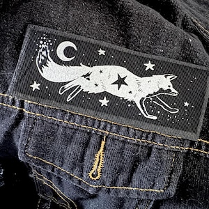 May include: A black rectangular patch featuring a white fox leaping across a starry night sky with a crescent moon. The patch is sewn onto a dark denim jacket, with a pocket and yellow stitching visible.