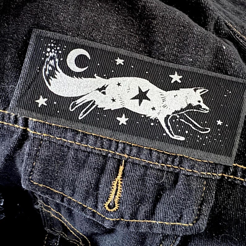 May include: A black rectangular patch featuring a white fox leaping across a starry night sky with a crescent moon. The patch is sewn onto a dark denim jacket, with a pocket and yellow stitching visible.