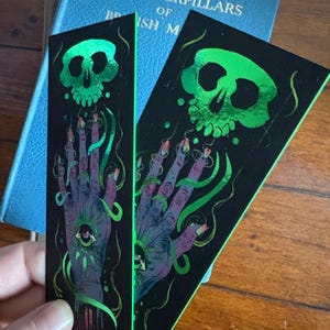 May include: Two black bookmarks with a green skull and hand design. The hand has an eye in the palm and long fingernails. Green, swirly lines connect the skull and hand. The bookmarks are next to a blue book.