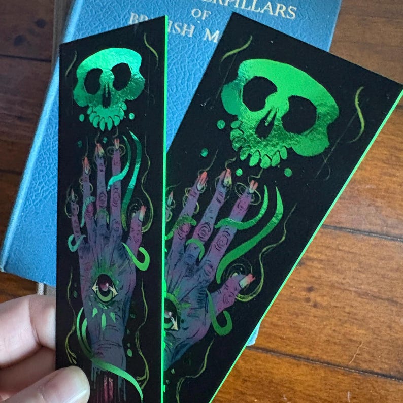 May include: Two black bookmarks with a green skull and hand design. The hand has an eye in the palm and long fingernails. Green, swirly lines connect the skull and hand. The bookmarks are next to a blue book.
