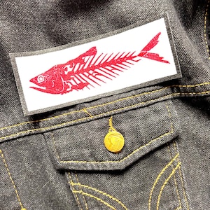 May include: A red fish skeleton patch on a dark denim jacket. The patch is rectangular with a white background and a detailed red fish bone design. The jacket has a gold button and yellow stitching.