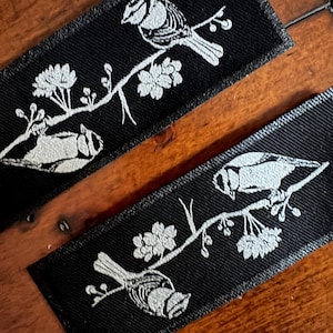 Könnte beinhalten: Zwei schwarze Stoffaufnäher mit einem weißen Vogel- und Blumenast-Design. Der Vogel sitzt auf einem Ast mit Blüten und Blättern. Die Aufnäher sind rechteckig und haben ein strukturiertes Aussehen.