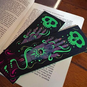May include: Two black bookmarks with a spooky design. Each bookmark features a green skull and a purple hand with a glowing green eye. The hand is adorned with long, pointed fingernails and green, swirling lines.