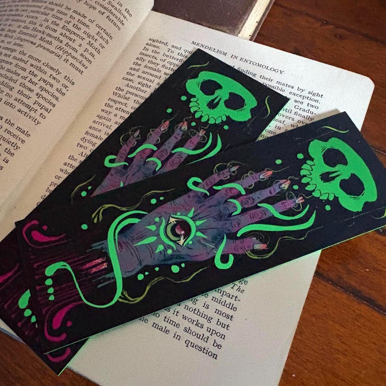 May include: Two black bookmarks with a spooky design. Each bookmark features a green skull and a purple hand with a glowing green eye. The hand is adorned with long, pointed fingernails and green, swirling lines.
