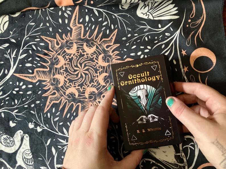 May include: A black and gold deck of cards called "Occult Ornithology" by K.S. Whittle. The cards feature a black and white illustration of a bird skull with a teal background.