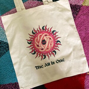 May include: A white canvas tote bag with a pink and black sun design and the text "The All is One".