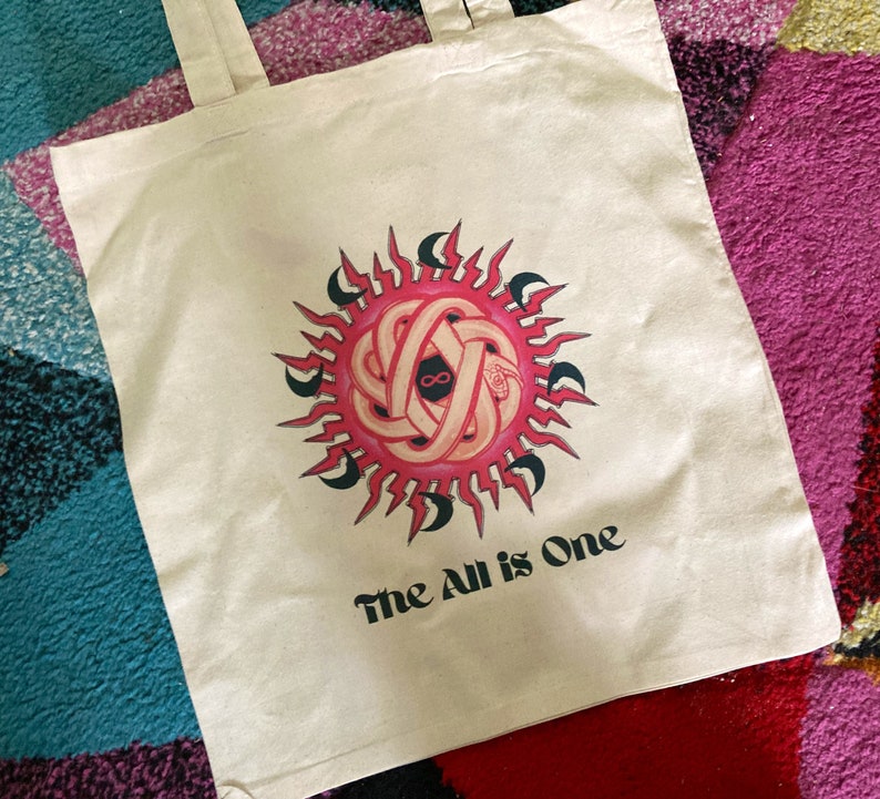 May include: A white canvas tote bag with a pink and black sun design and the text "The All is One".