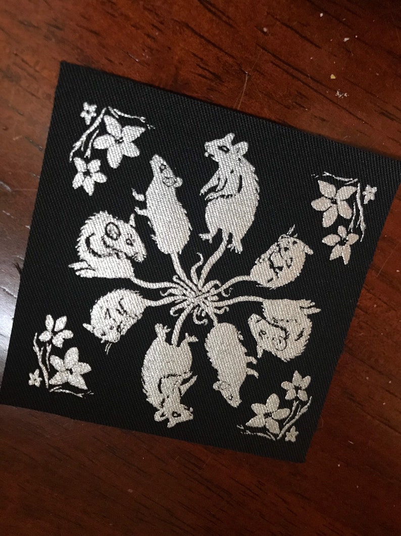 May include: A black fabric patch with a white design of eight mice in a circle, with their tails intertwined in the center. The mice are facing outward, with their ears and whiskers visible. There are small white flowers around the edge of the patch.