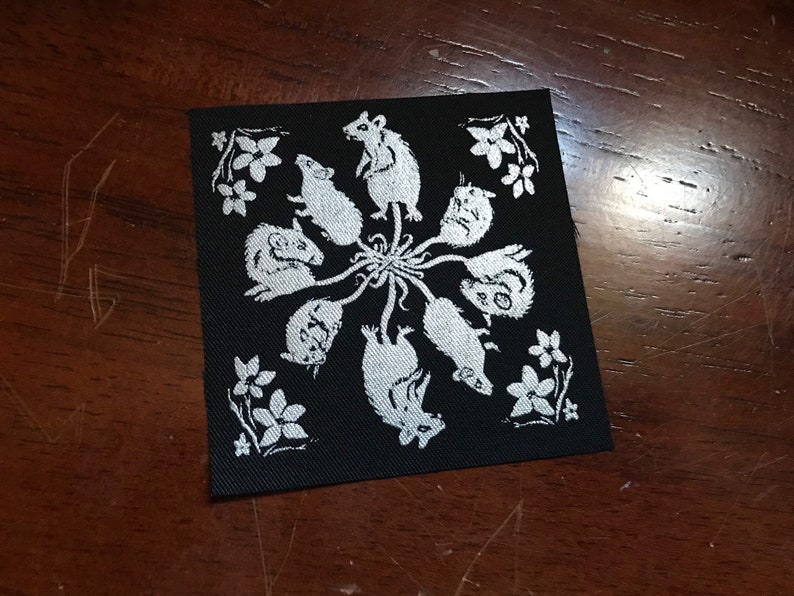 May include: A black fabric patch with a white design of eight mice in a circle around a central point. The mice are facing outward and have long tails that extend to the center. The background is black and the mice are white.