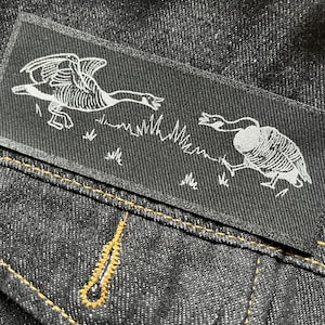 Könnte beinhalten: Eine schwarze Jeansjacke mit einem weißen gestickten Aufnäher von zwei sich gegenüberstehenden Gänsen. Der Aufnäher ist auf die vordere Tasche der Jacke genäht.