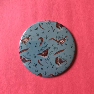 May include: A round blue mirror with a pattern of brown pheasants and arrows. The pheasants have text written on them, including "Strut", "Pretty Boy", "Babaanki", and "So Handsome".