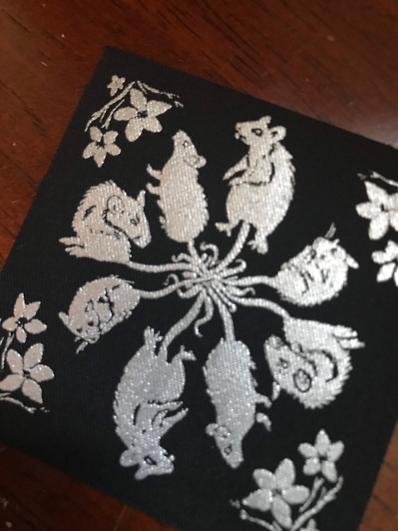 May include: A black fabric patch with a silver design of nine mice in a circle, with flowers around the edge.