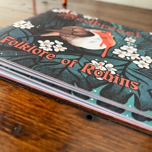 May include: A closed book titled "Folklore of Robins" with a robin redbreast illustration on the cover. The cover features a green, blue, and white floral pattern.