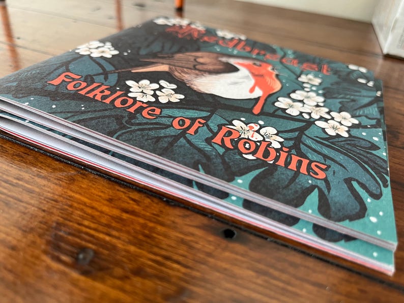 May include: A closed book titled "Folklore of Robins" with a robin redbreast illustration on the cover. The cover features a green, blue, and white floral pattern.