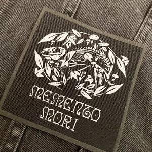 Pode incluir: Patch quadrado preto com uma ilustração branca de uma caveira, folhas e cogumelos. As palavras "Memento Mori" são impressas em branco, com letras góticas. O patch está sobre um fundo de tecido cinza escuro.