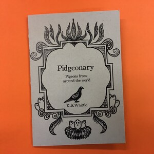 以下が含まれることがあります： 装飾的なフレームとハトのイラストが描かれた「Pidgeonary」というタイトルの小さな本。本の表紙は、黒インクのライトグレーです。タイトルはハトのイラストの上にあり、「世界中のハト」というテキストがあります。
