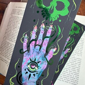May include: A dark gray bookmark with a neon green skull and a hand with an eye in the palm. The hand is in shades of blue, purple, and pink with long, pointed nails. Green and black accents complete the design.