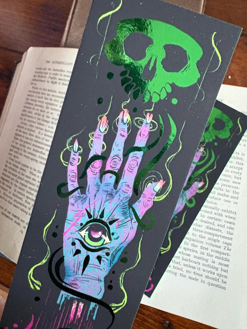 May include: A dark gray bookmark with a neon green skull and a hand with an eye in the palm. The hand is in shades of blue, purple, and pink with long, pointed nails. Green and black accents complete the design.