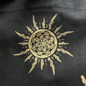 May include: A black tote bag with a gold sun design. The sun has a swirling pattern inside and is surrounded by rays.