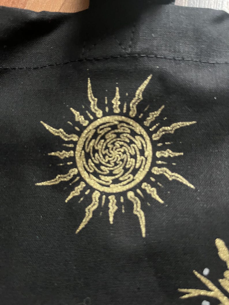 May include: A black tote bag with a gold sun design. The sun has a swirling pattern inside and is surrounded by rays.