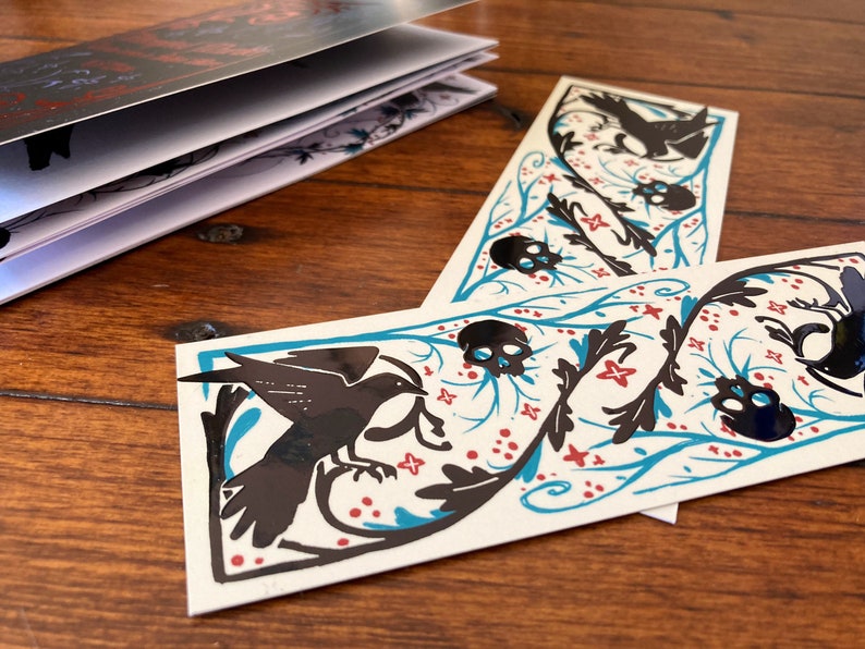 May include: Two black and white bookmarks with a black bird, skulls, and floral designs on a teal background. The bookmarks have red accents.
