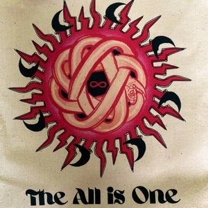 May include: A beige tote bag with a red and black sun design and the text "The All is One" printed in black below the sun.
