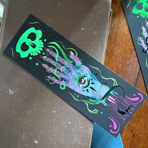 May include: Black bookmark with a neon green skull and a hand with an eye in the palm. The hand is purple and blue with long, pointed nails. Green and pink accents complete the design. The bookmark is resting on a brown book.