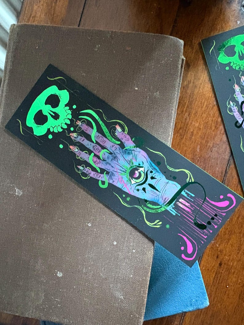 May include: Black bookmark with a neon green skull and a hand with an eye in the palm. The hand is purple and blue with long, pointed nails. Green and pink accents complete the design. The bookmark is resting on a brown book.