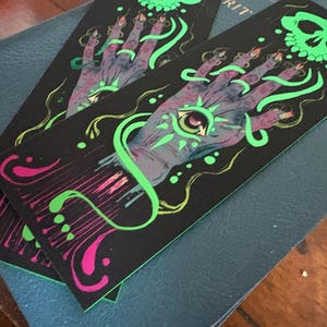 May include: Two black bookmarks with a mystical design. Each features a purple hand with a glowing green eye, green swirls, and a green skull. The design is accented with pink and yellow details.