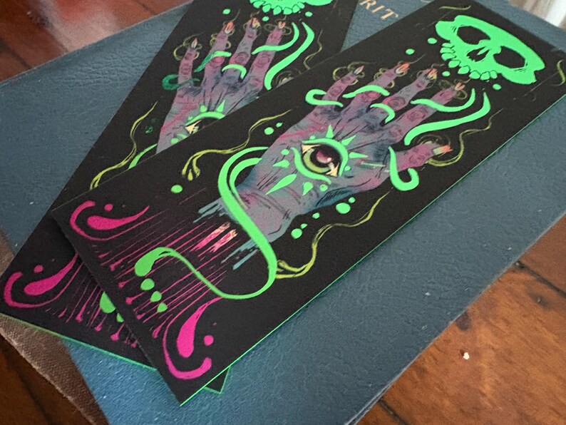 May include: Two black bookmarks with a mystical design. Each features a purple hand with a glowing green eye, green swirls, and a green skull. The design is accented with pink and yellow details.