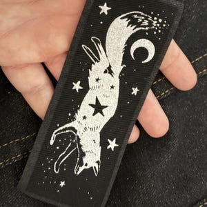 May include: Black rectangular patch featuring a white illustration of a fox hanging upside down, surrounded by stars and a crescent moon. The fox has a star on its chest. The patch is held by a hand.