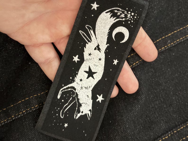 May include: Black rectangular patch featuring a white illustration of a fox hanging upside down, surrounded by stars and a crescent moon. The fox has a star on its chest. The patch is held by a hand.