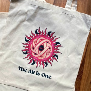May include: A white tote bag with a pink and black sun design and the text "The All is One".