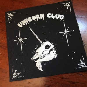 May include: Black square patch with white text and illustration. The text reads "UNICORN CLUB" above a unicorn skull with a long horn. White stars and dots are in the background.