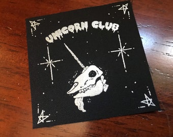 May include: Black square patch with white text and illustration. The text reads "UNICORN CLUB" above a unicorn skull with a long horn. White stars and dots are in the background.