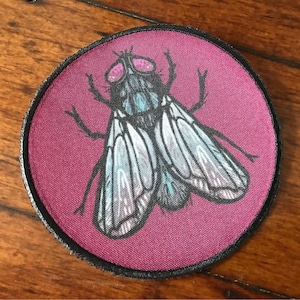 May include: A round, burgundy fabric patch with a black border featuring a detailed illustration of a fly with iridescent wings.