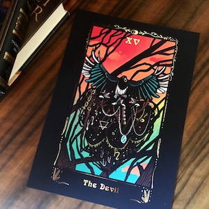 May include: A black tarot card print featuring a magpie with teal and black wings, set against a sunset-colored sky. The card is labeled "The Devil" with gold accents and the Roman numeral XV.