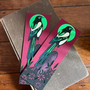 以下が含まれることがあります： 2つのブックマーク。カササギのイラストが特徴です。カササギは、黒、白、緑の詳細で描かれ、緑の円のあるバーガンディの背景に設定されています。ブックマークの下部には花の模様があります。